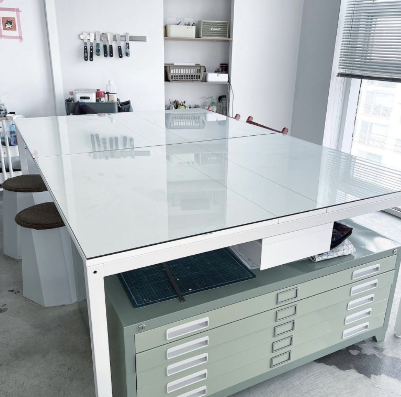 A white studio workstation with a glass top and a green metal storage cabinet with drawers in an art workshop.