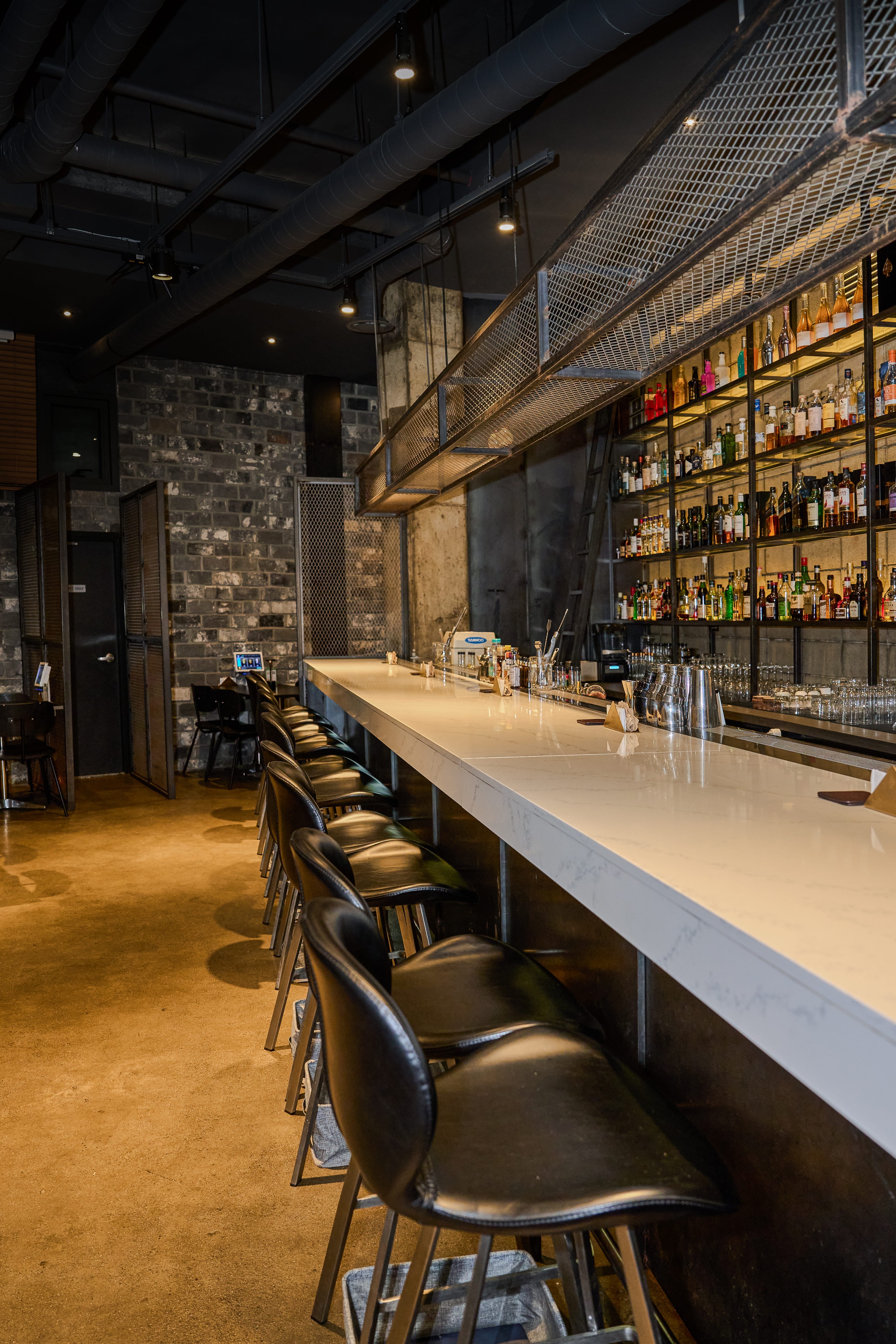 Interior of a modern bar with a long marble-topped counter and rows of liquor bottles on back-lit shelves.