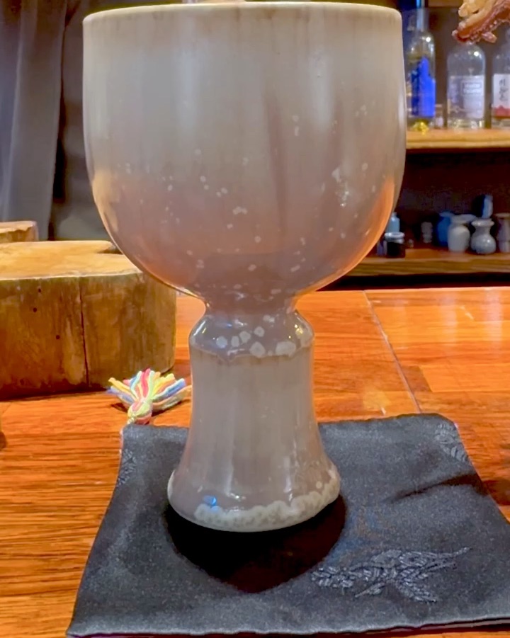 A beige ceramic goblet sits on a dark coaster on a wooden bar counter.