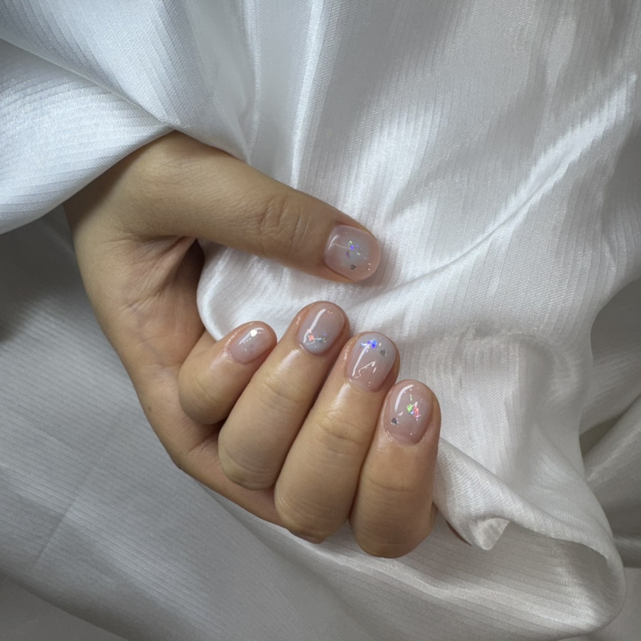 Close-up of a hand with clean, short nail art featuring iridescent glitter accents against a white silk fabric background