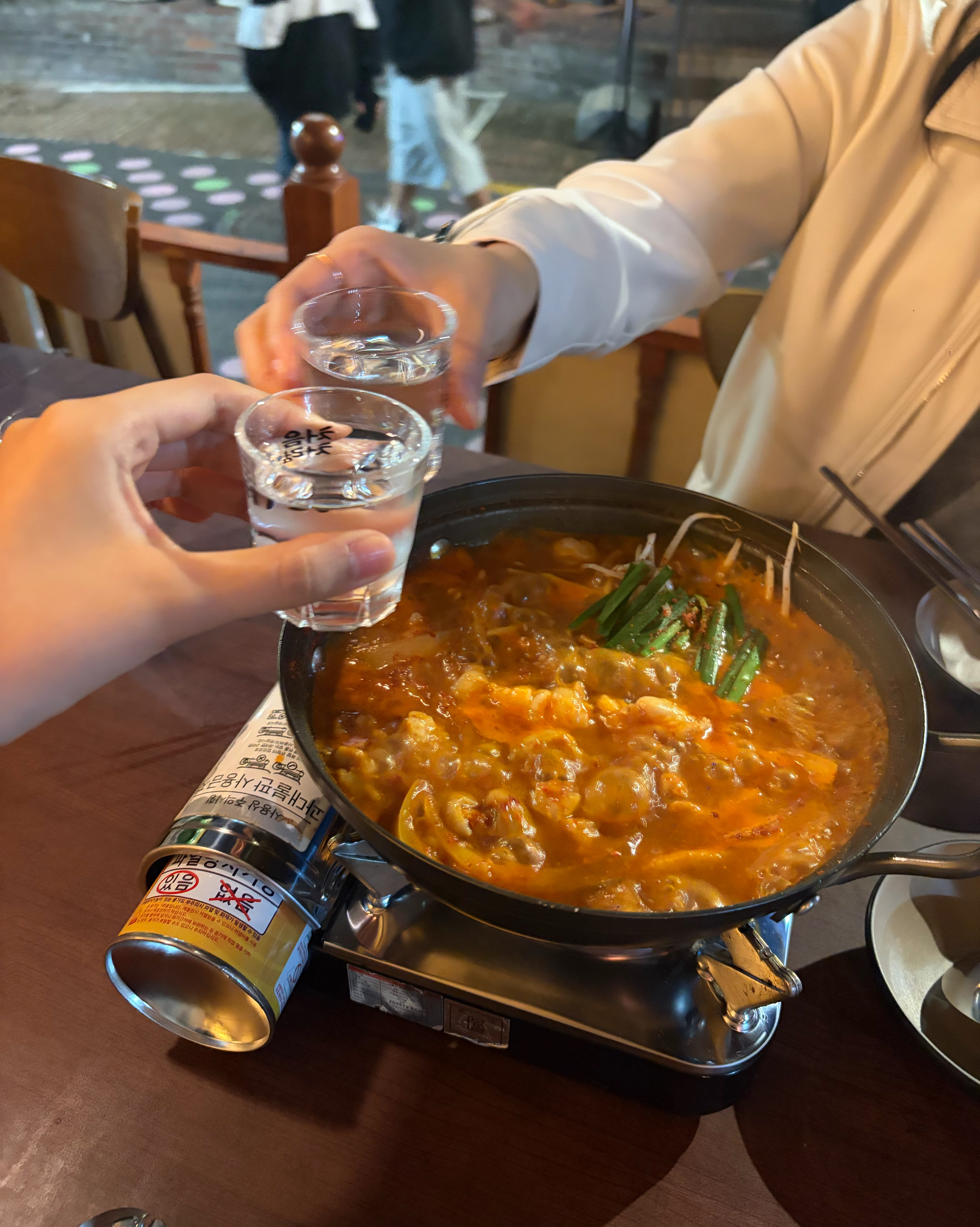 Two guests toasting soju glasses over a simmering spicy stew pot at Daejeop