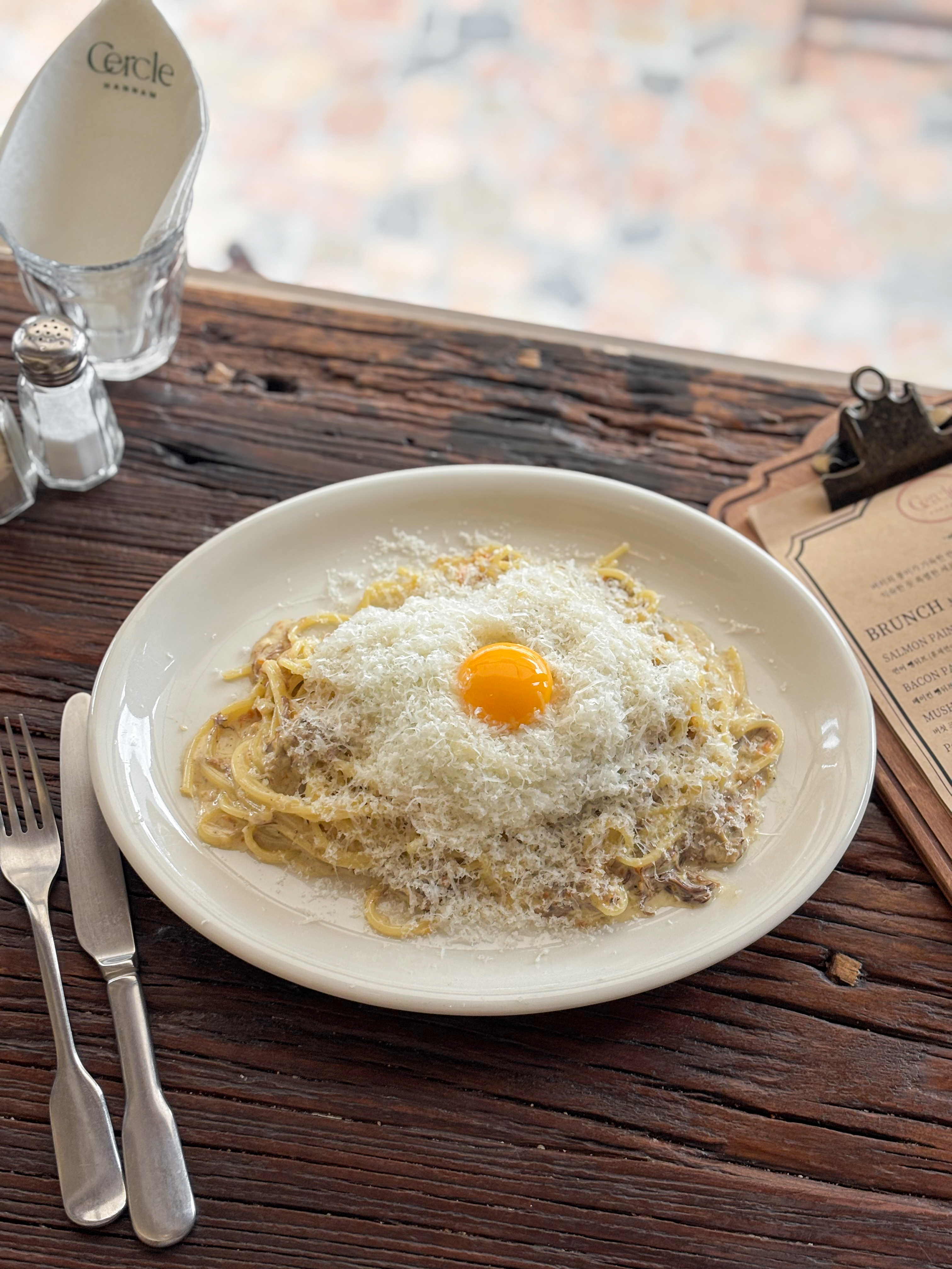 A plate of pasta topped with a raw egg yolk and grated cheese on a rustic wooden table at Cercle Hannam.