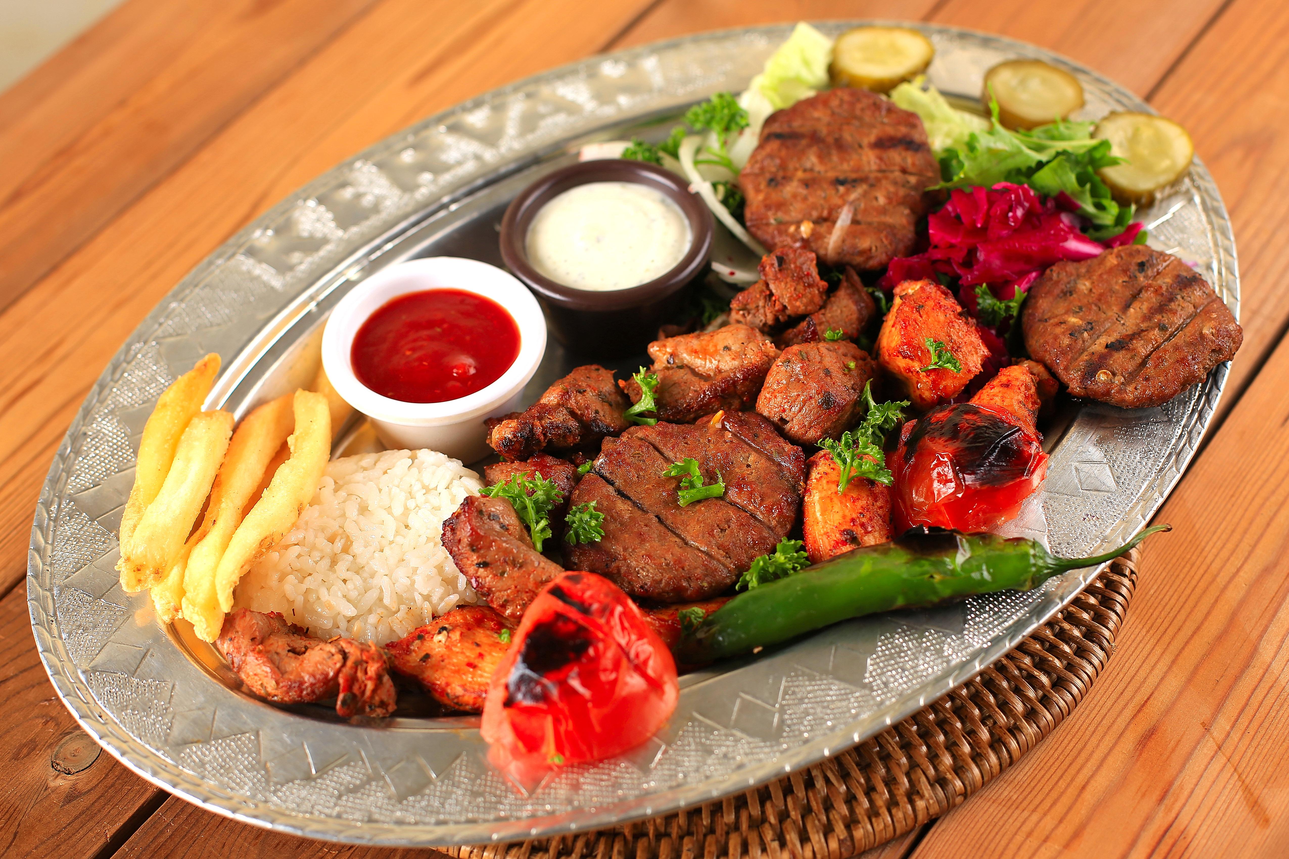 A metal platter featuring a mixed grill meal with meats, rice, french fries, vegetables, and two sauces.