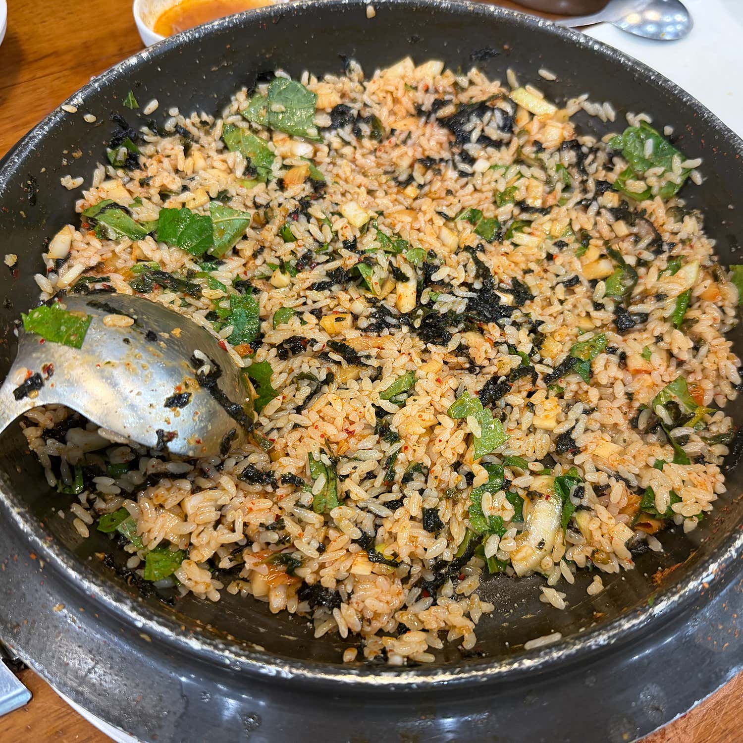 A close-up overhead shot of Korean fried rice in a black metal pan with a stainless steel spoon.