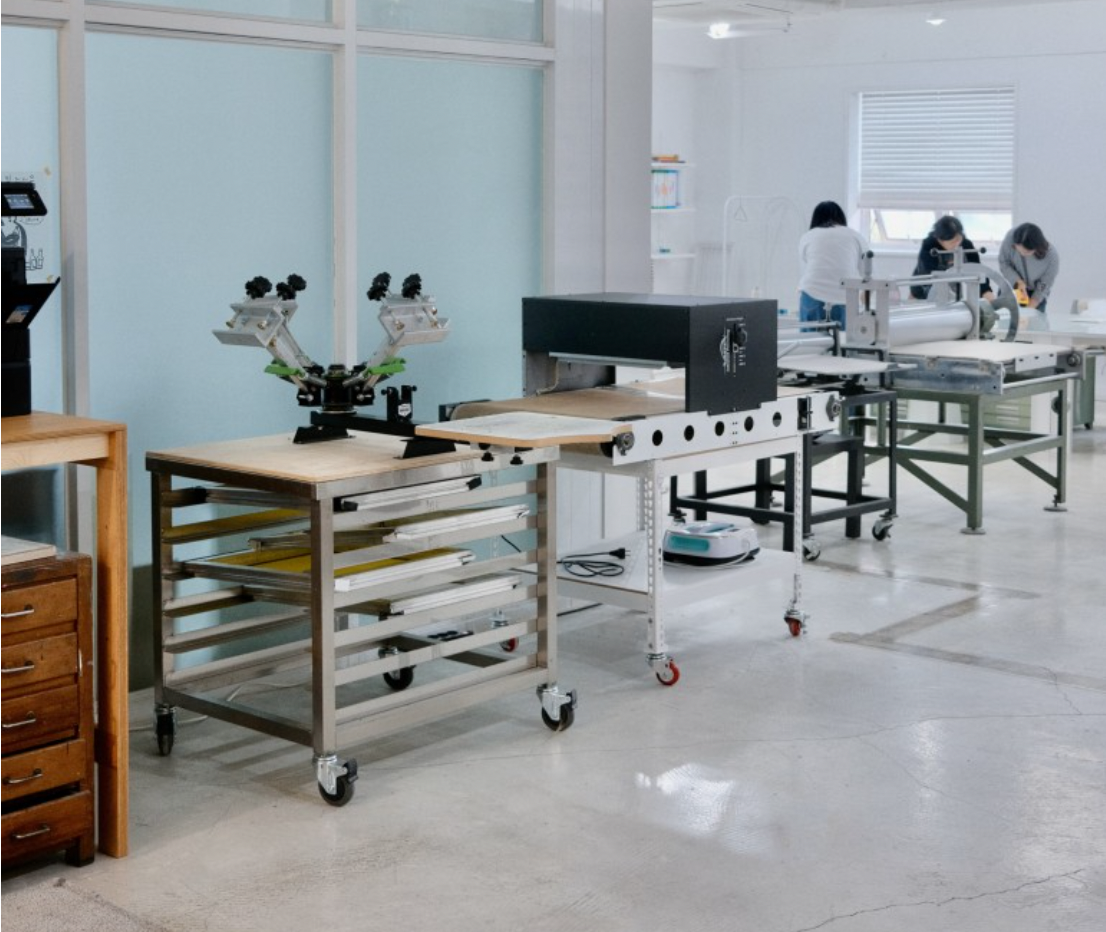 Interior of an art studio with screen printing equipment and people working in the background.