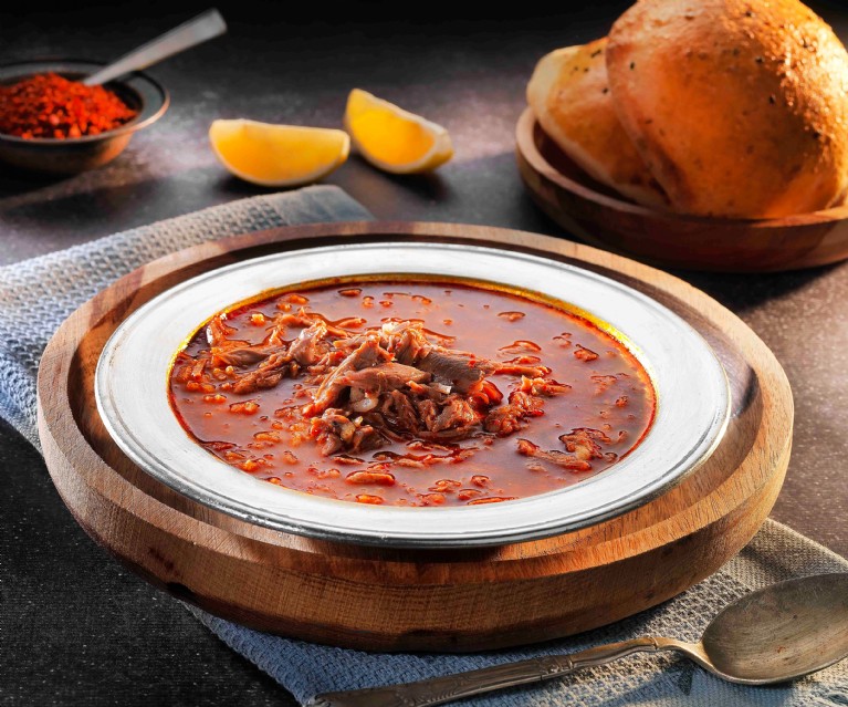 A metal bowl of meat soup served on a wooden board with bread and lemon wedges.