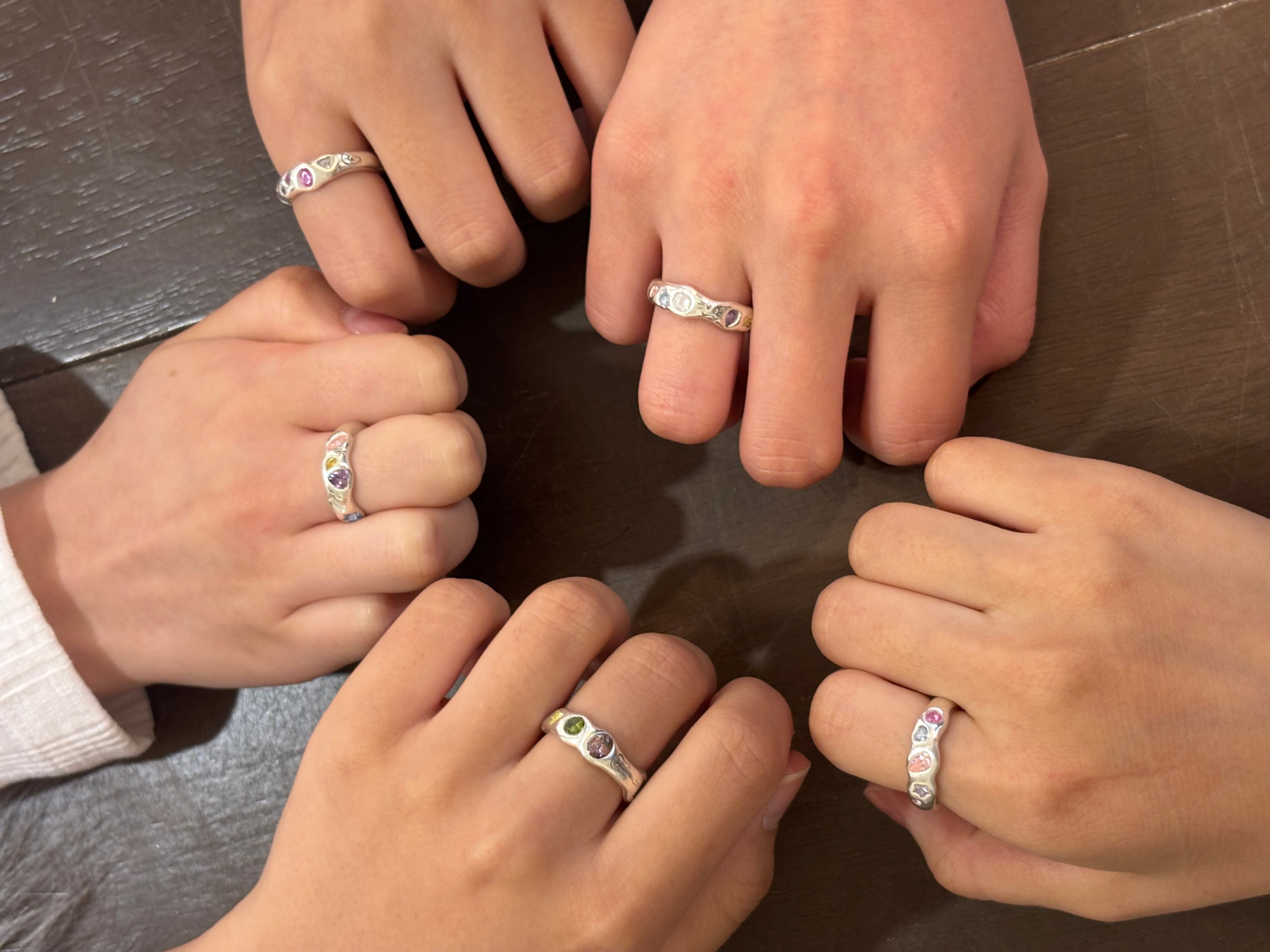 Close-up of four hands wearing silver rings with small colorful gemstones, arranged in a circle on a dark wooden table.