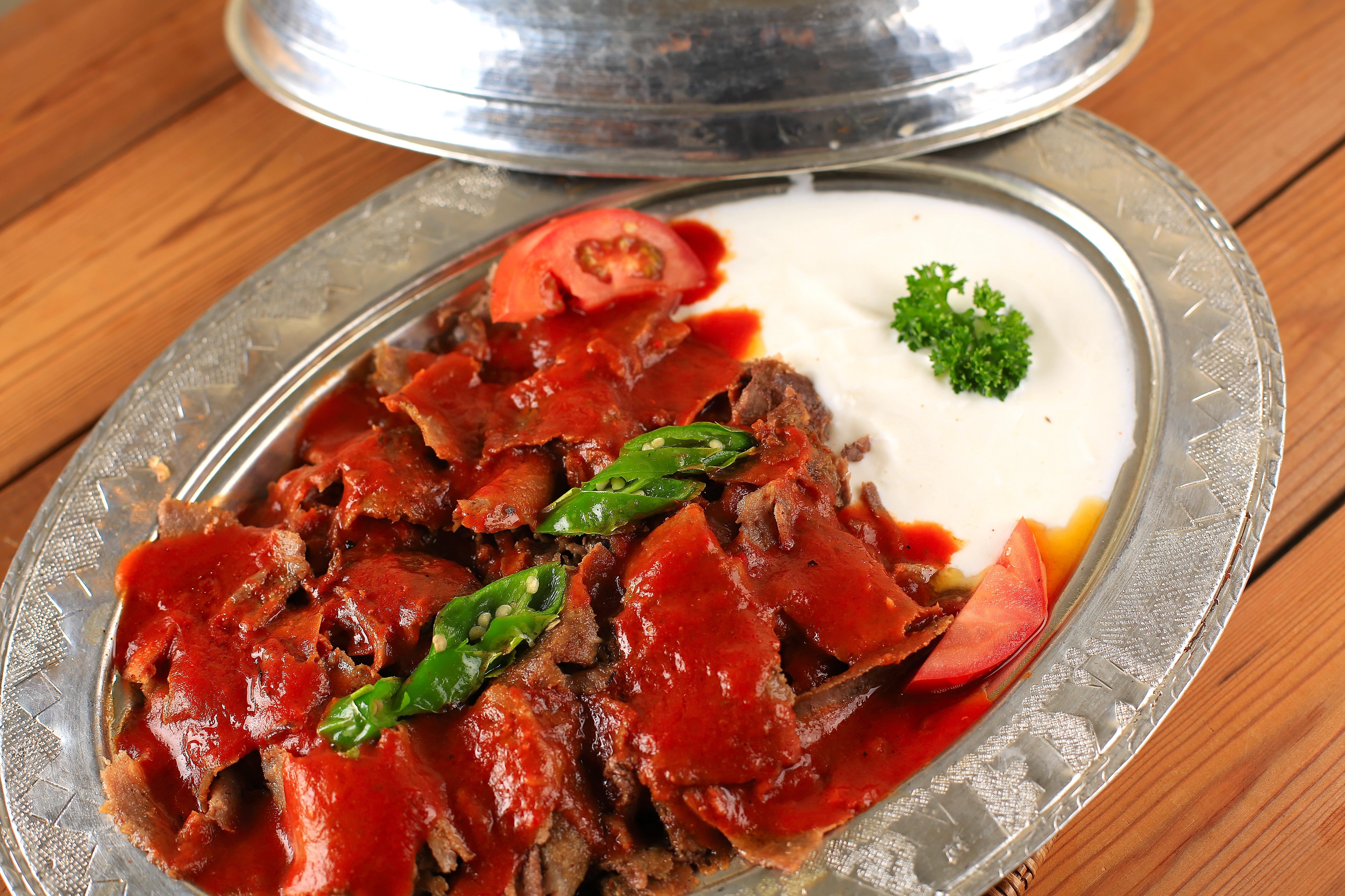 A metal serving platter containing sliced meat in red sauce with white yogurt and fresh garnishes.