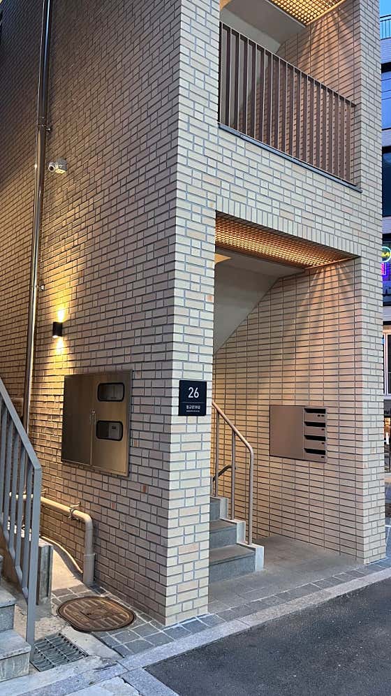 Exterior view of the modern beige brick building entrance at 26 Donggyo-ro 38-gil with warm wall lights.
