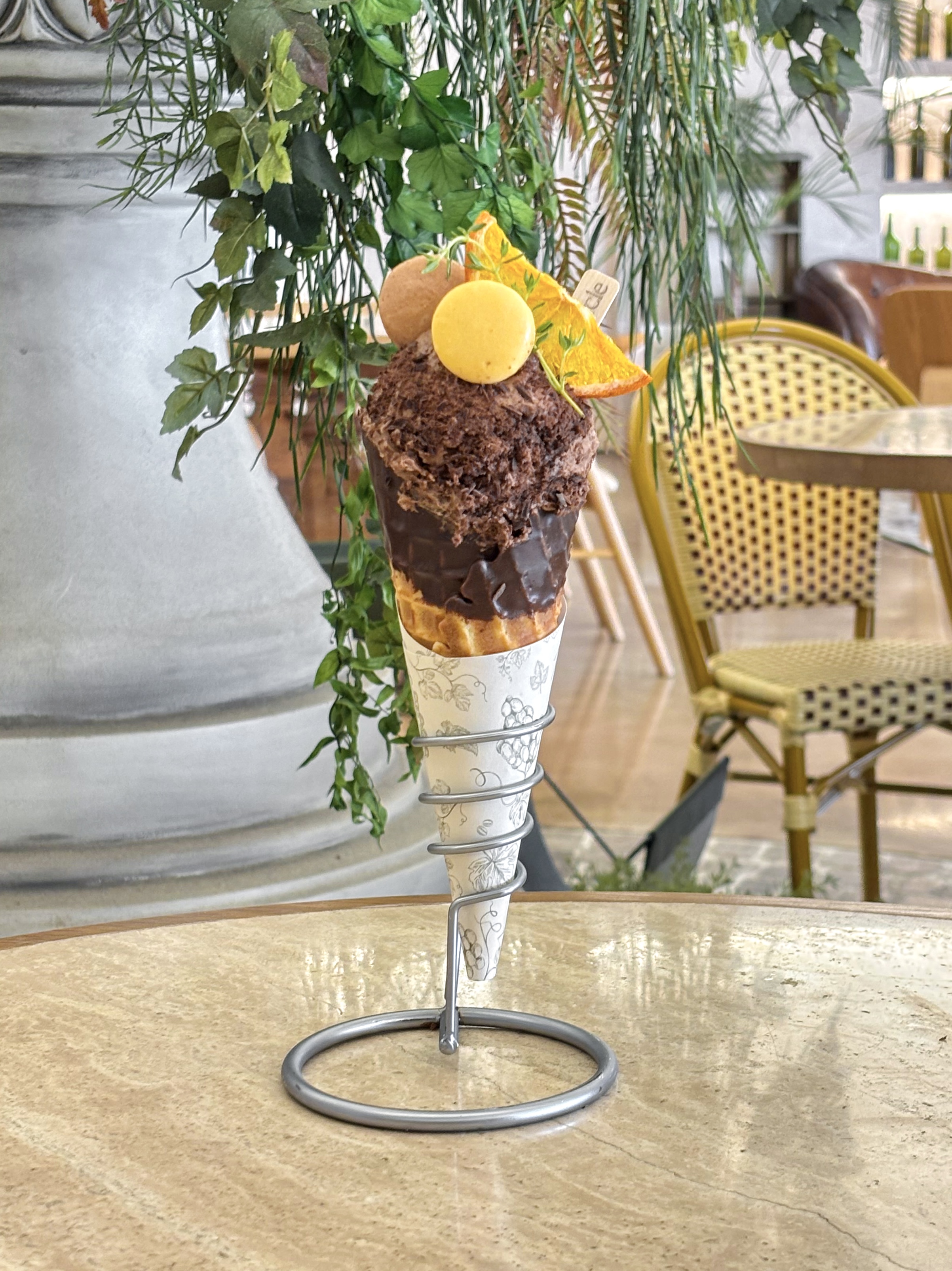A chocolate ice cream cone topped with a macaron and dried orange slice, displayed in a metal stand on a marble cafe table.