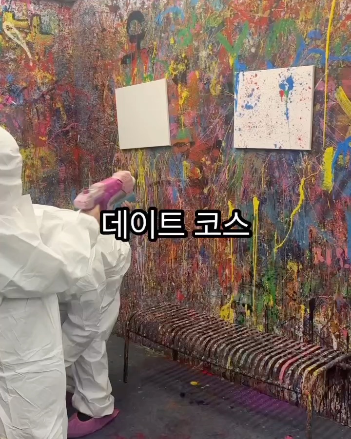 Person in white protective suit using a paint gun in a studio with walls covered in colorful paint splatters