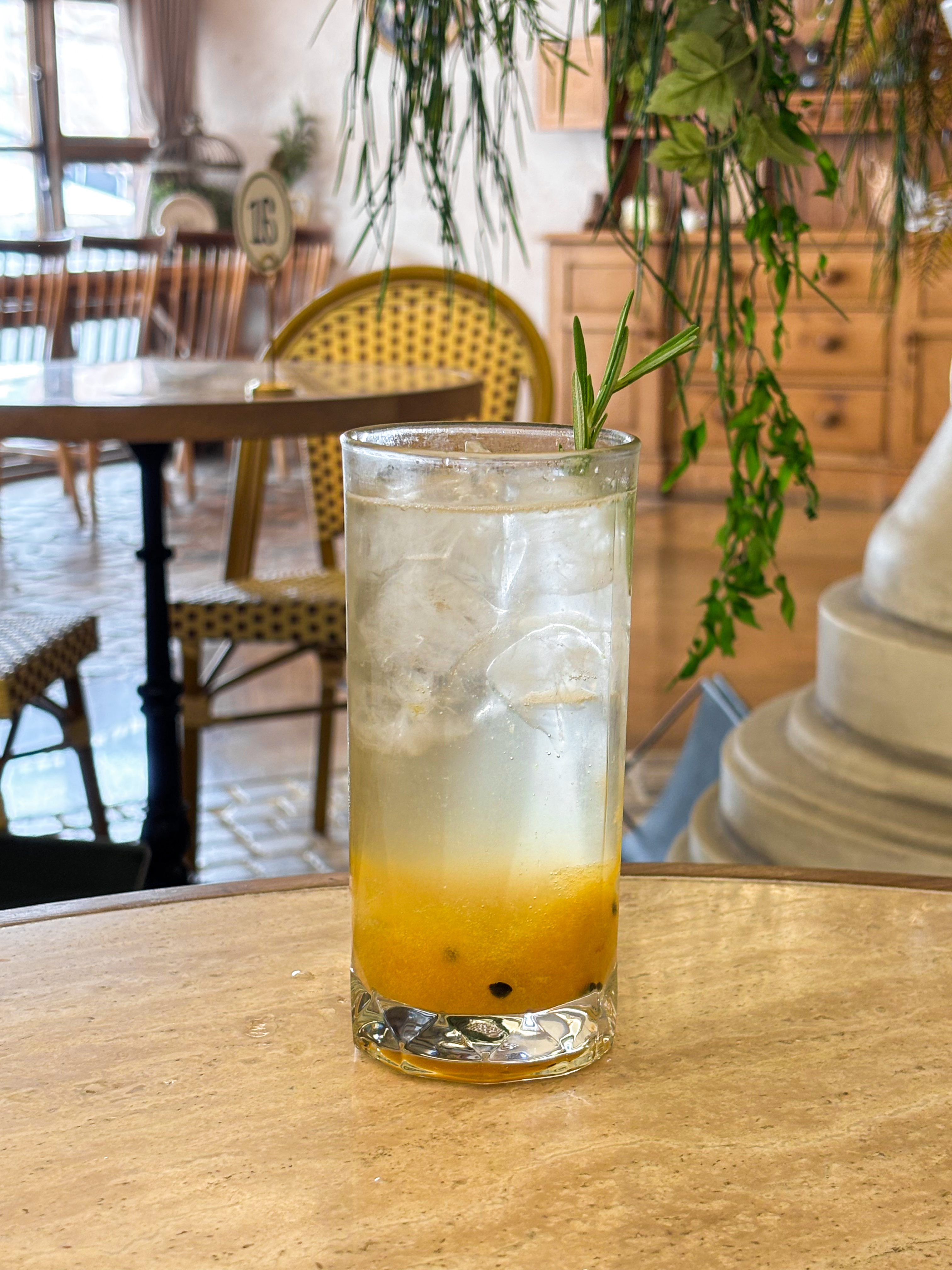 A clear glass of iced passion fruit drink with a rosemary sprig on a cafe table at Cercle Hannam.