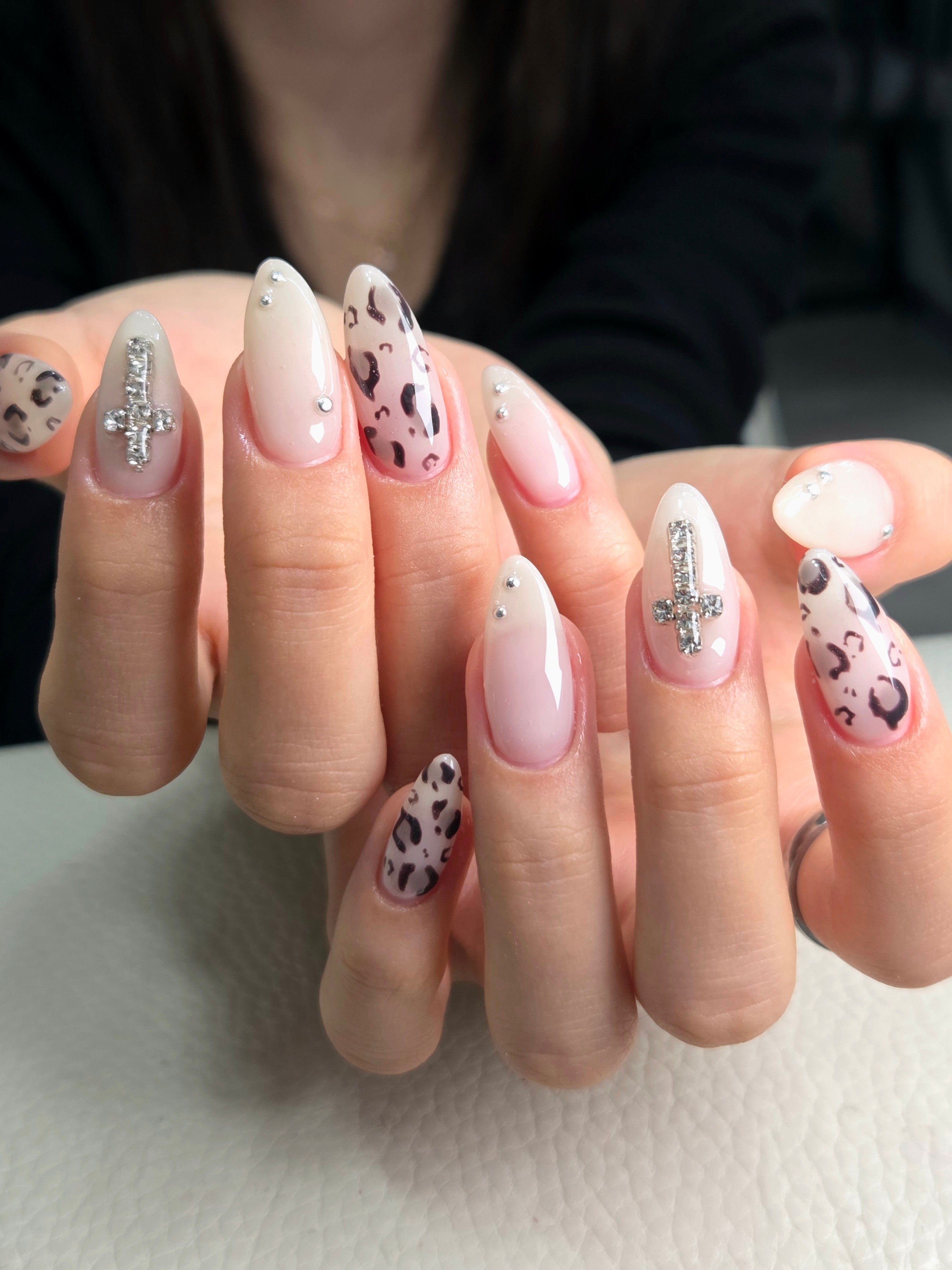 Close-up of almond-shaped nails featuring leopard print, rhinestone cross charms, and silver studs on a light gradient base.