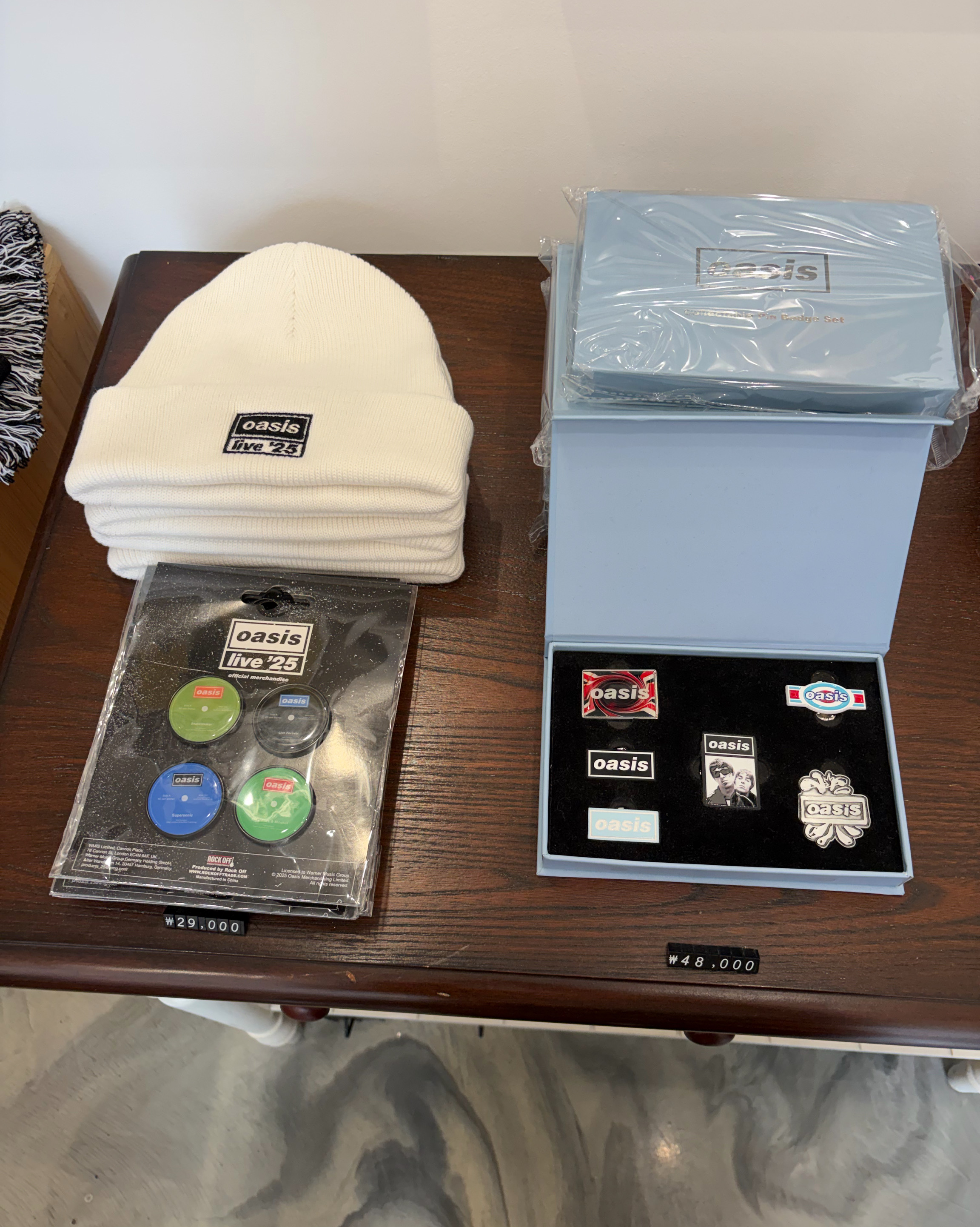 Oasis band merchandise including beanies, button pins, and a badge set displayed on a dark wooden table