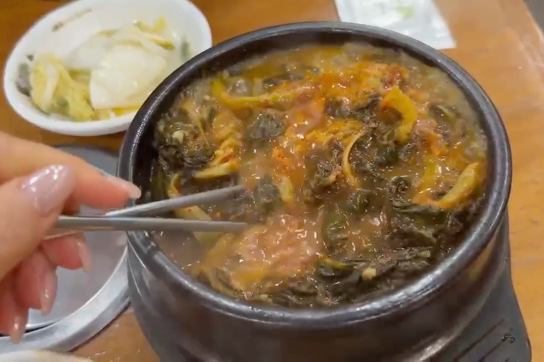 A steaming hot bowl of Korean gamjatang soup with greens and a side of kimchi.