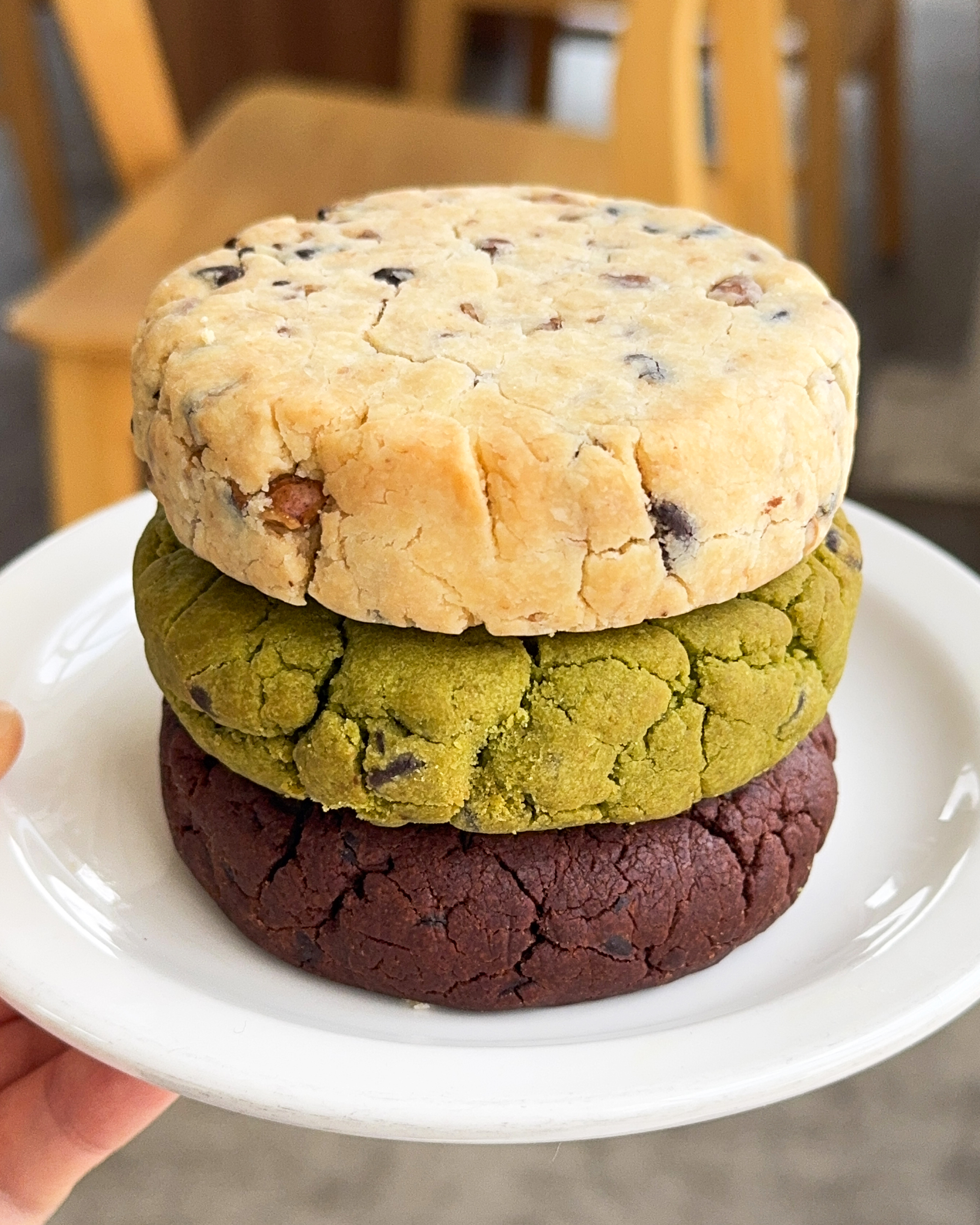 A stack of three thick, round, textured cookies—one beige, one green, and one dark brown—resting on a white plate.