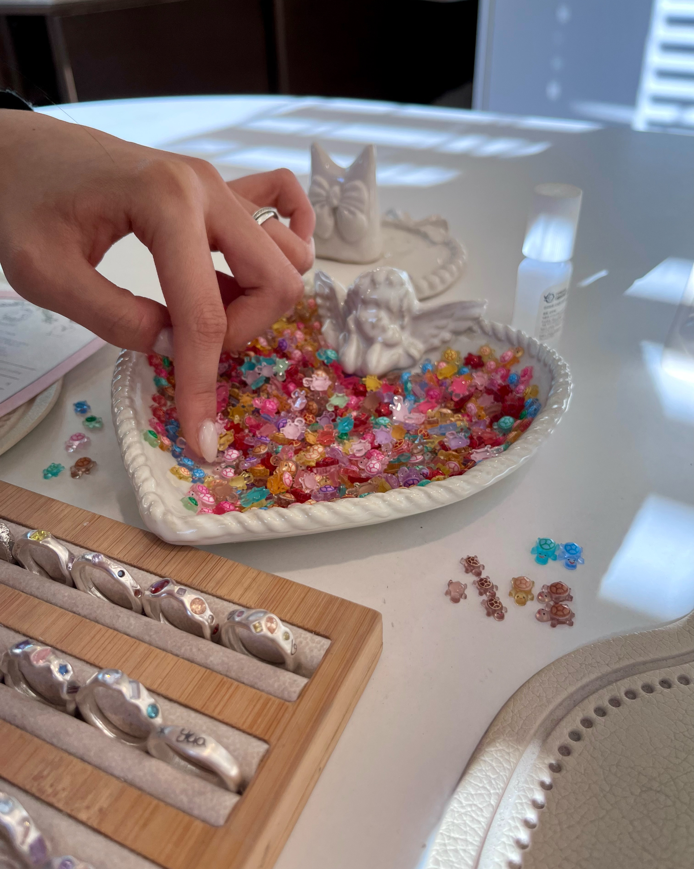 A person selecting small colorful turtle-shaped beads from a white heart-shaped dish on a white table.