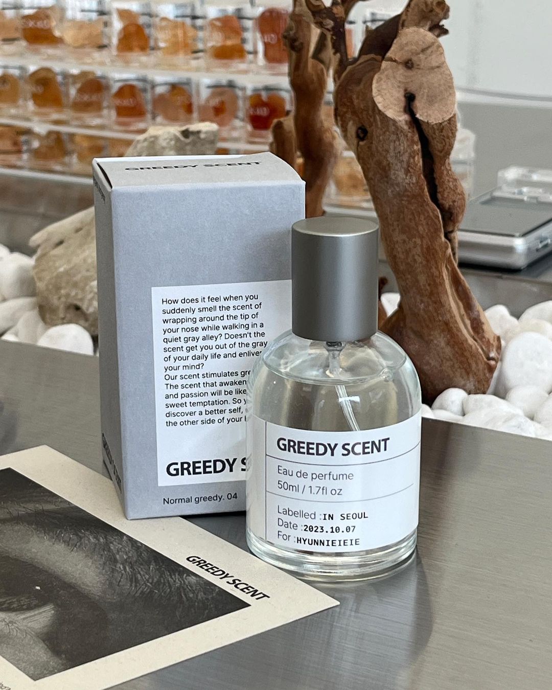 A bottle of Greedy Scent perfume and its packaging box displayed on a metallic studio surface