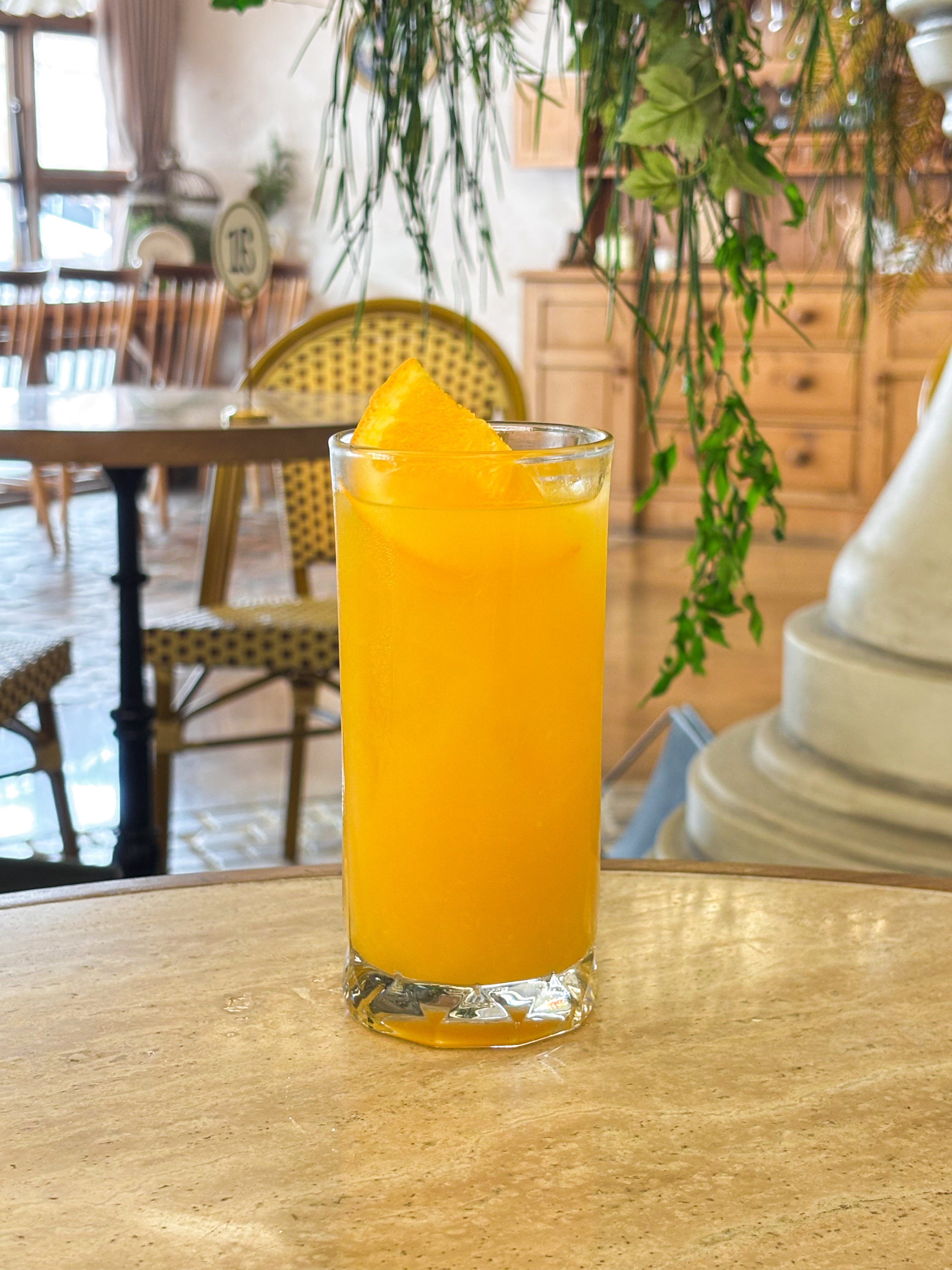 A glass of orange juice with a slice of orange on a cafe table, with a blurred cafe interior background.