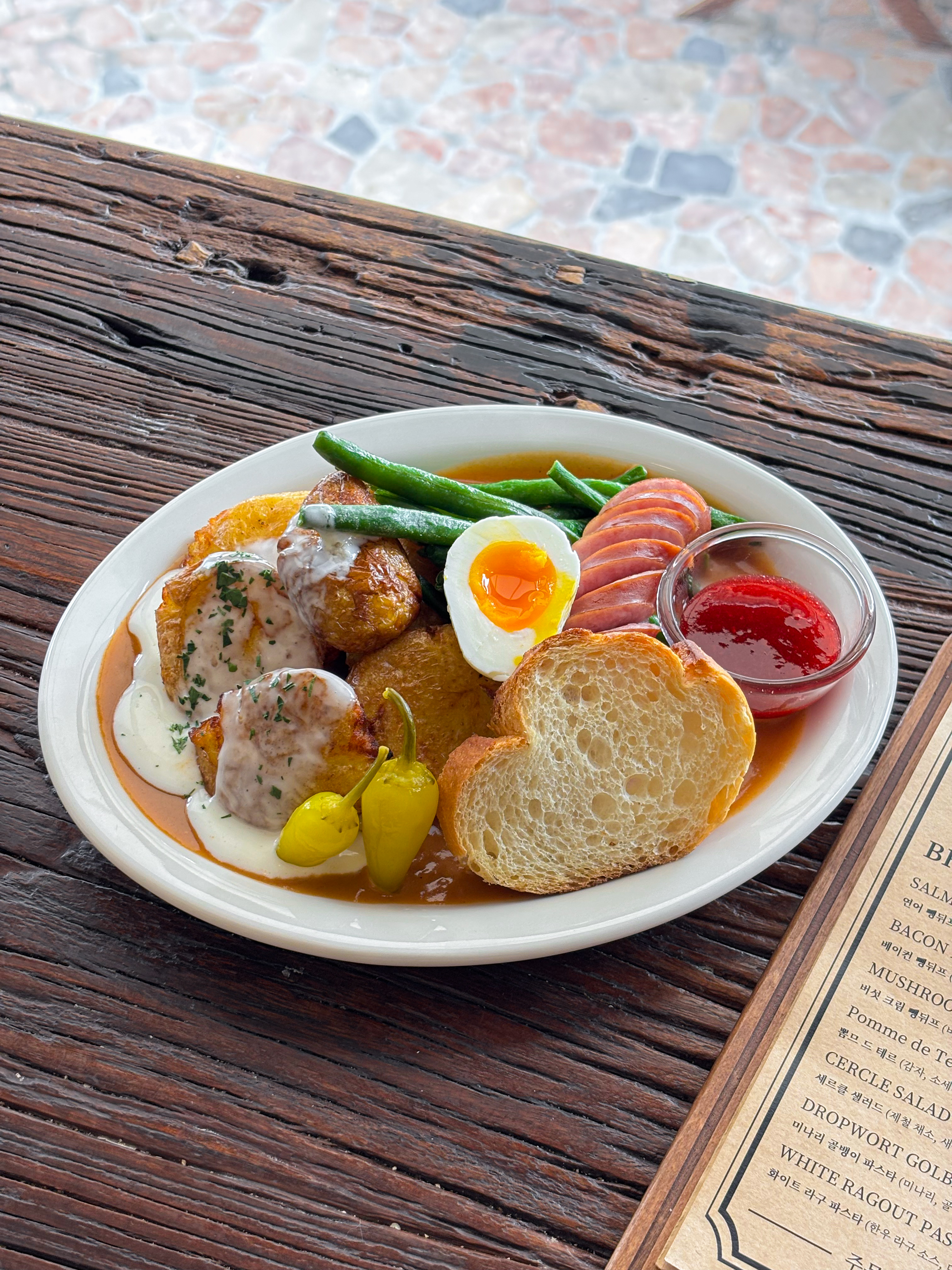 A plate of brunch food featuring roasted potatoes, green beans, sliced sausage, a soft-boiled egg, and bread with jam.