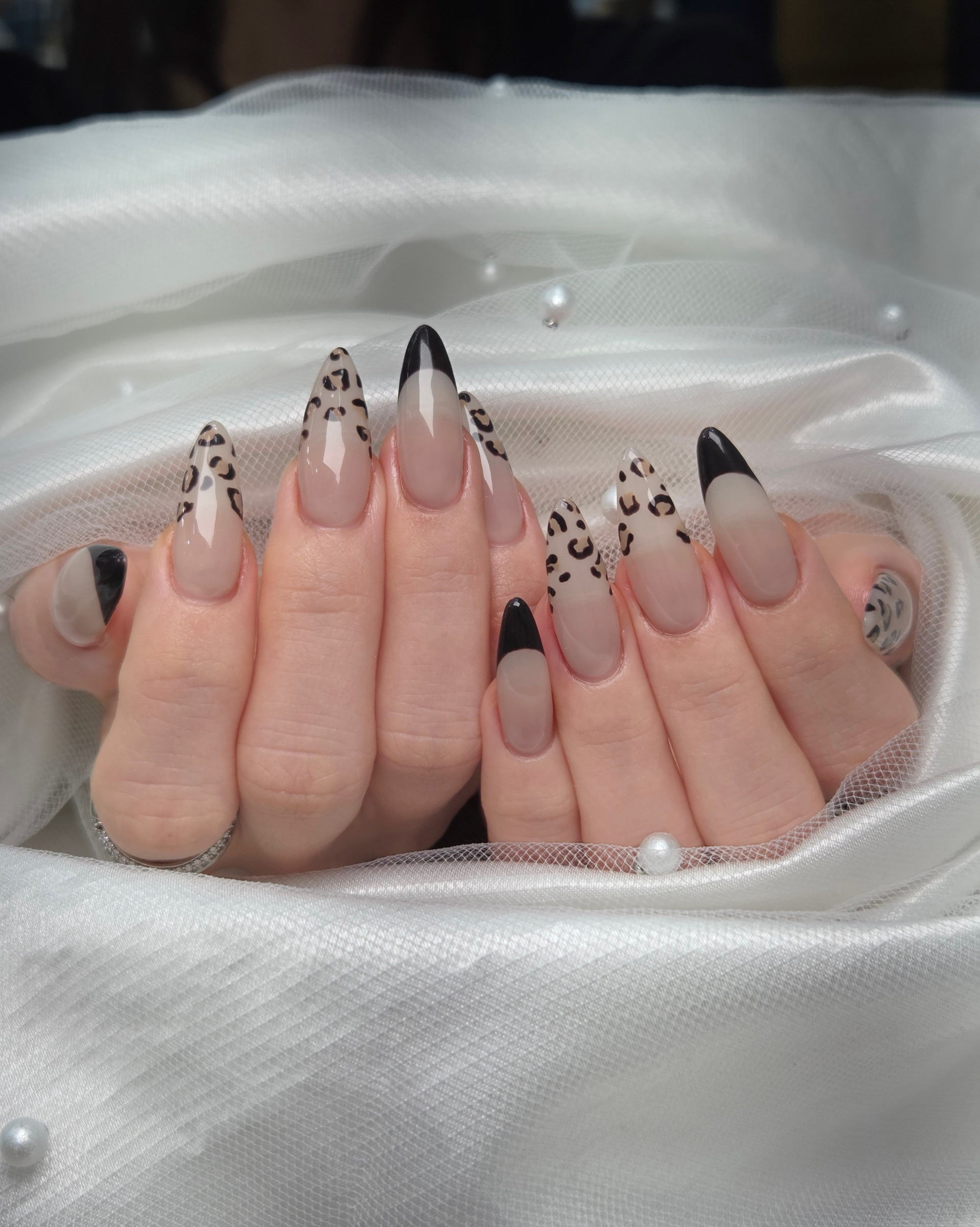 Close-up of almond-shaped fingernails with leopard print and black tip nail art designs, resting on white fabric with pearls