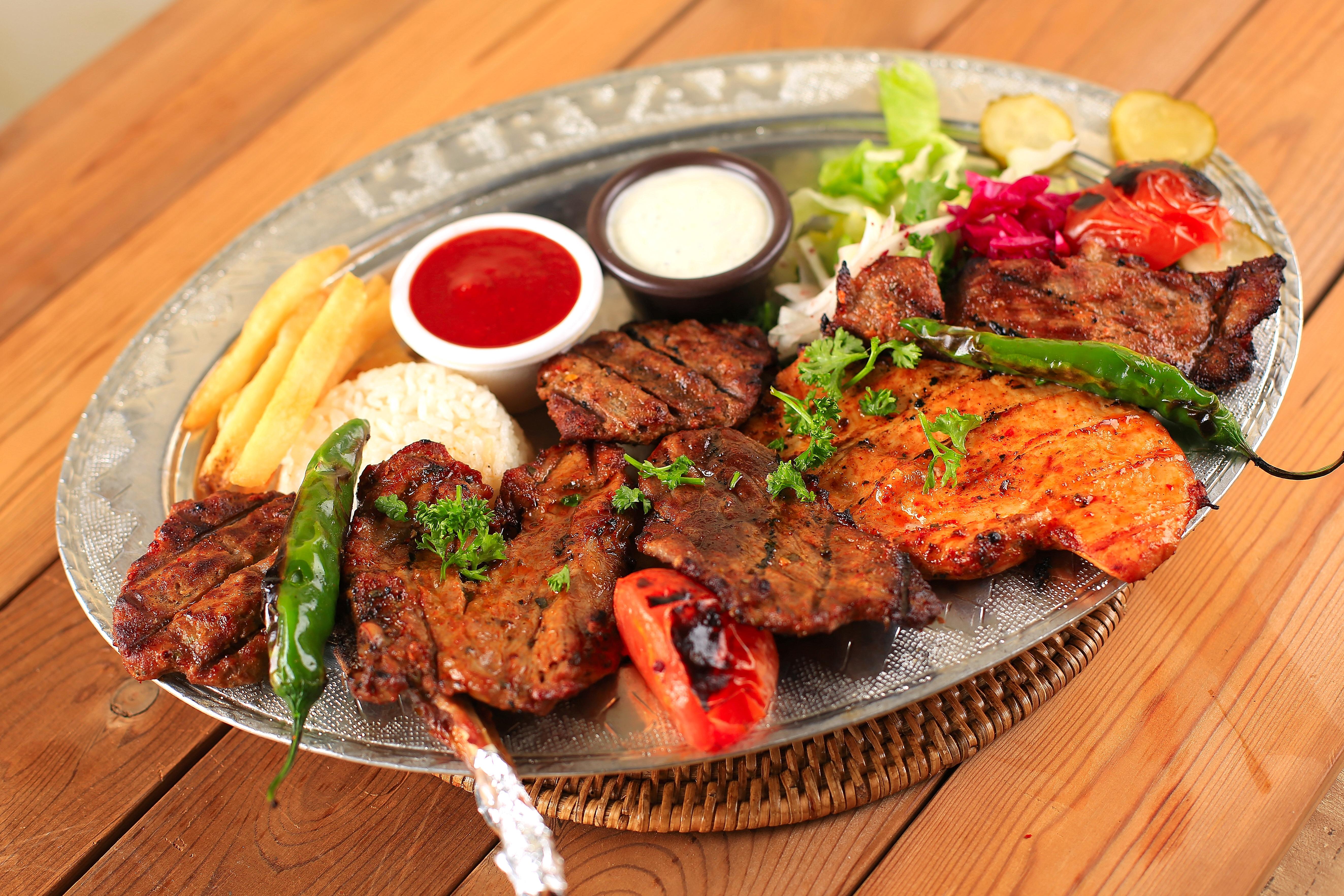 A mixed grill platter with various grilled meats, rice, french fries, and dipping sauces on a silver tray.