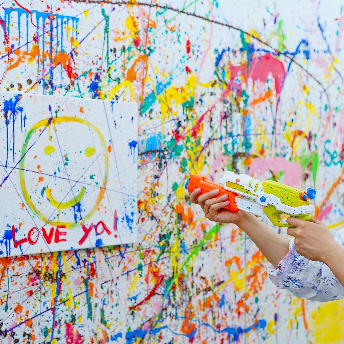 A person holding a toy paint gun in front of a white canvas splattered with colorful paint