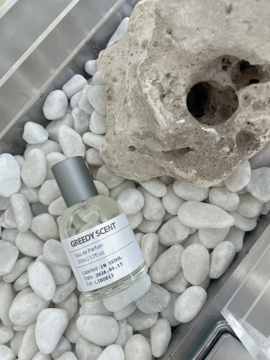 A bottle of Greedy Scent perfume placed among white decorative pebbles next to a textured stone