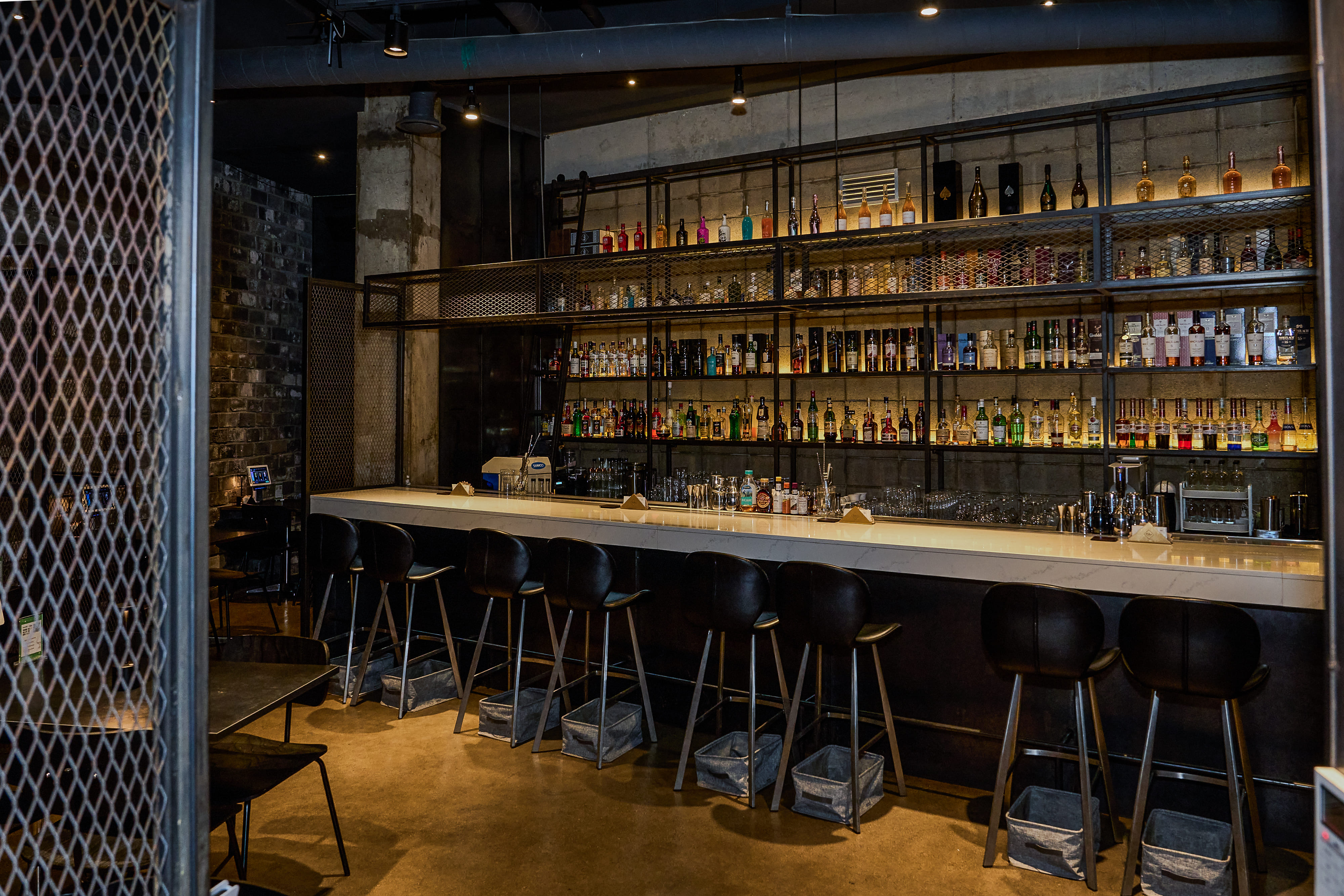 A dimly lit industrial style bar counter with rows of bottles on shelves and modern black bar stools.