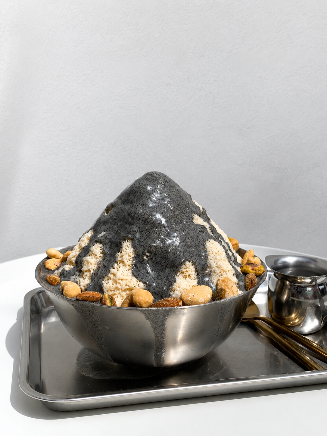A metal bowl filled with shaved ice topped with black sesame sauce and nuts on a tray.