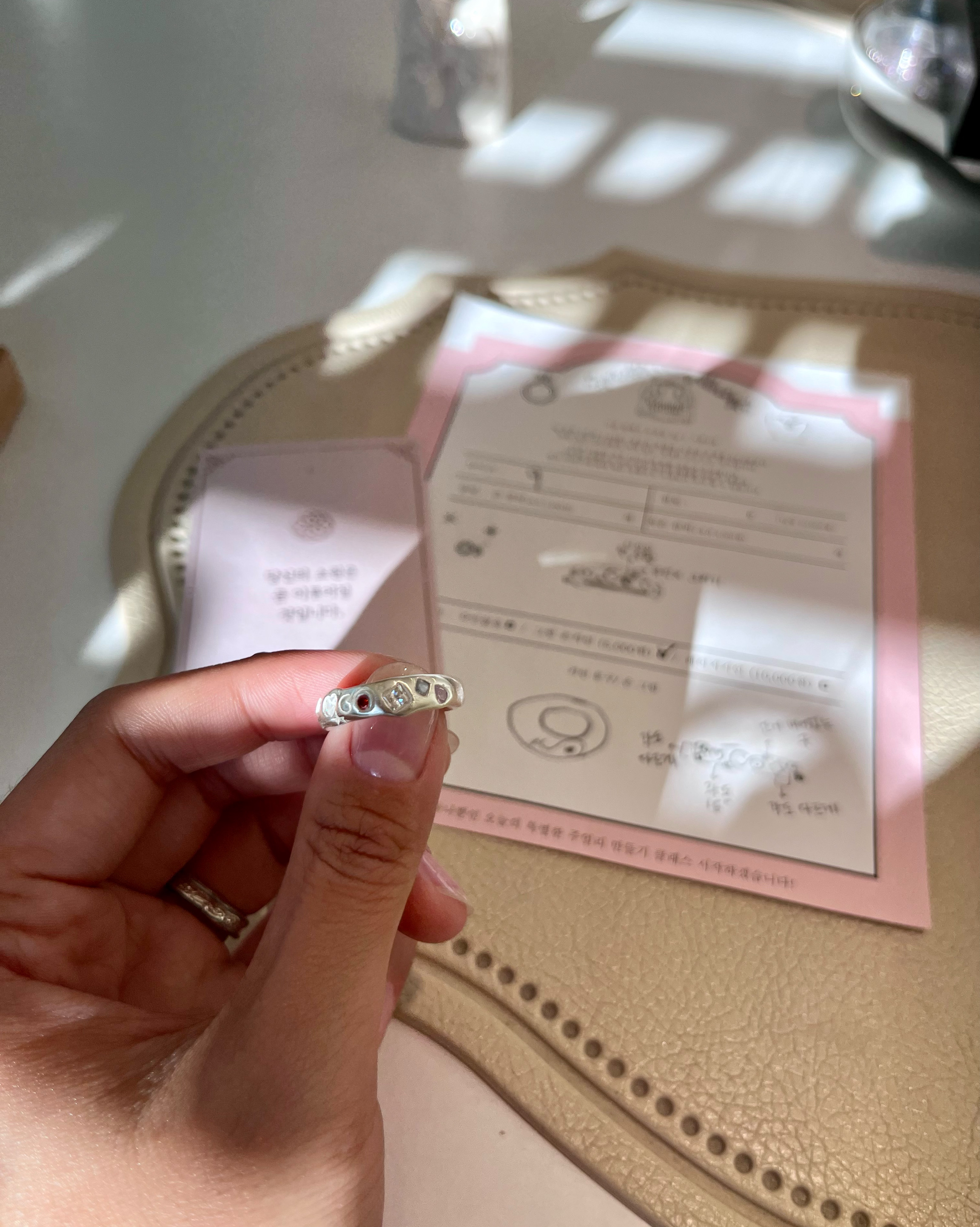 A hand holding a custom silver ring with small colorful stones over a desk with workshop paperwork.