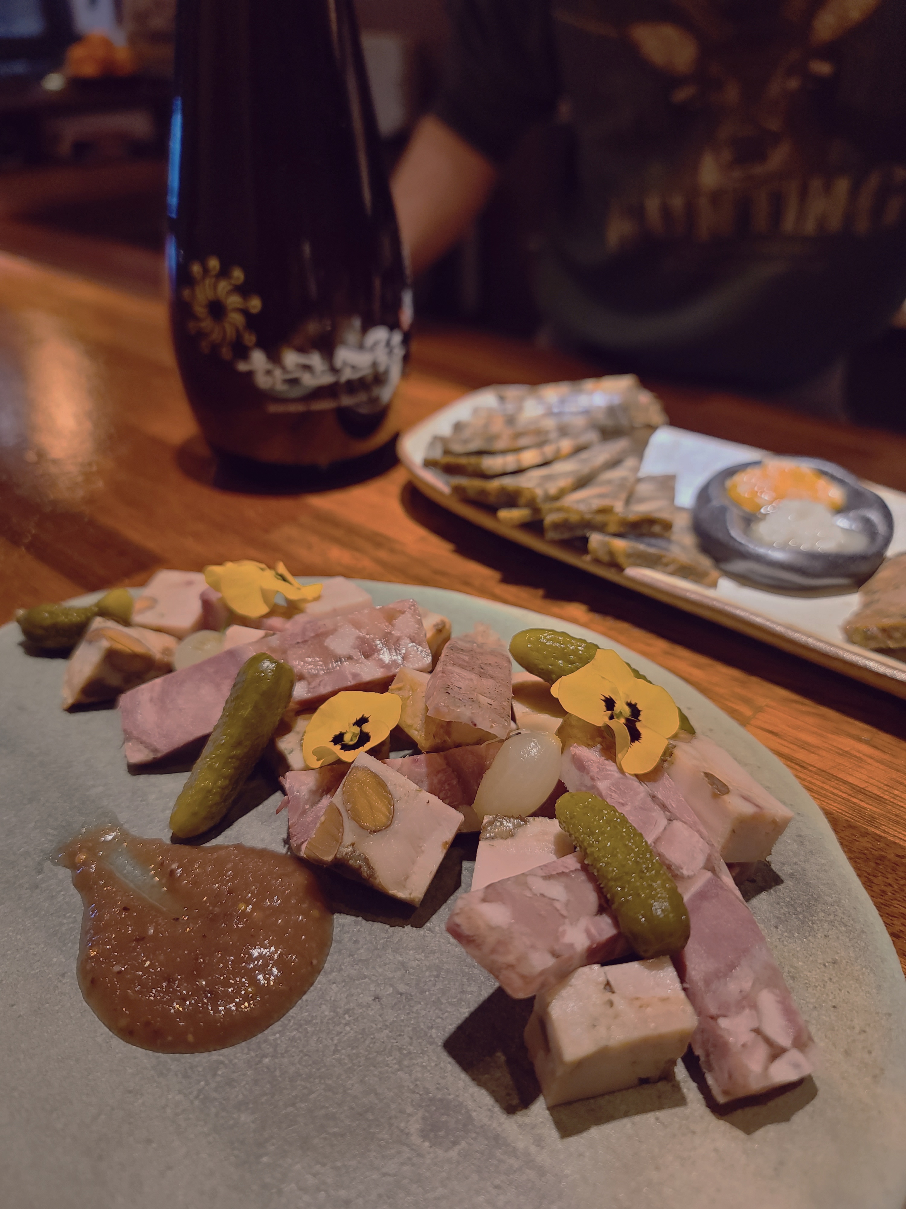 A plate of terrine with gherkins, mustard, and edible flowers on a wooden table at Namsan Alcohol Club.
