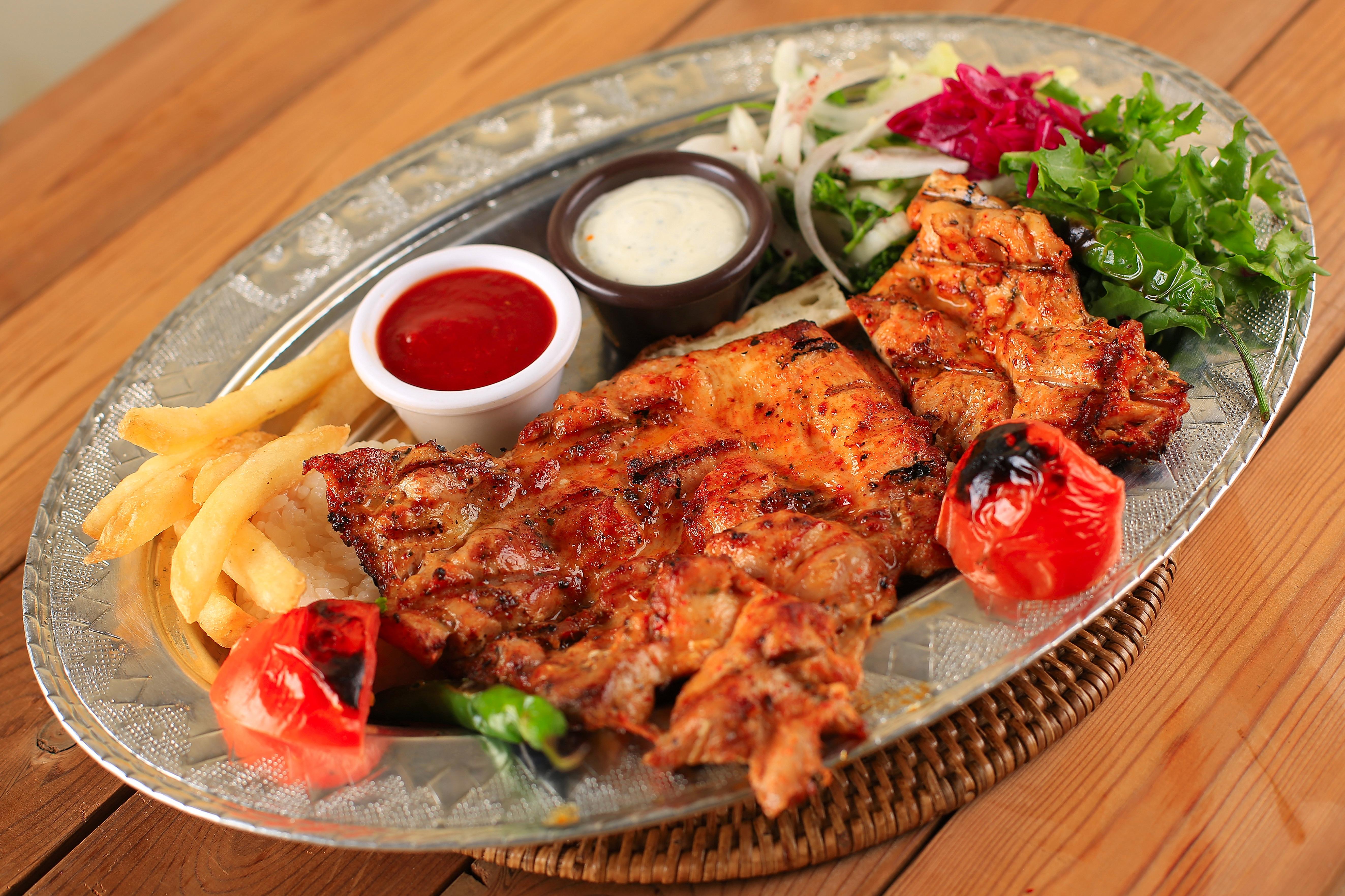 A platter featuring grilled chicken, fries, fresh salad, and two dipping sauces on a metallic serving tray.