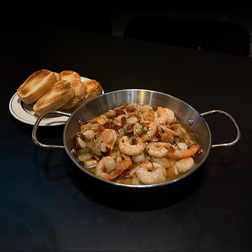 A metal skillet filled with cooked shrimp in oil with herbs served with toasted bread slices on a dark table.
