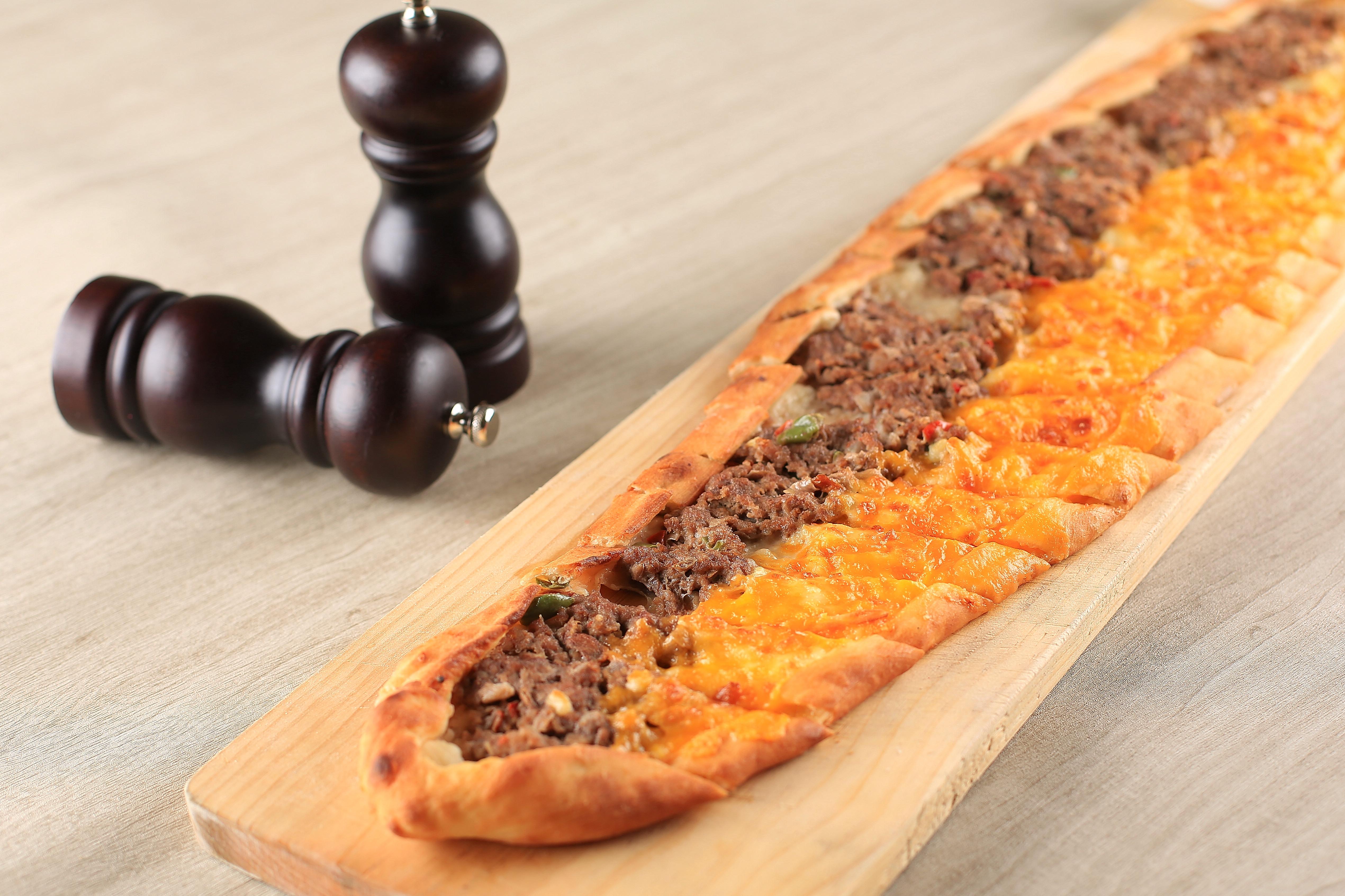 A long, boat-shaped Turkish pide flatbread with meat and cheese on a wooden board next to two pepper mills.