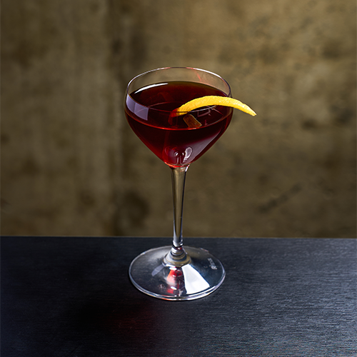 A deep red cocktail in a stemmed glass with a citrus peel twist, set against a blurred textured wall.
