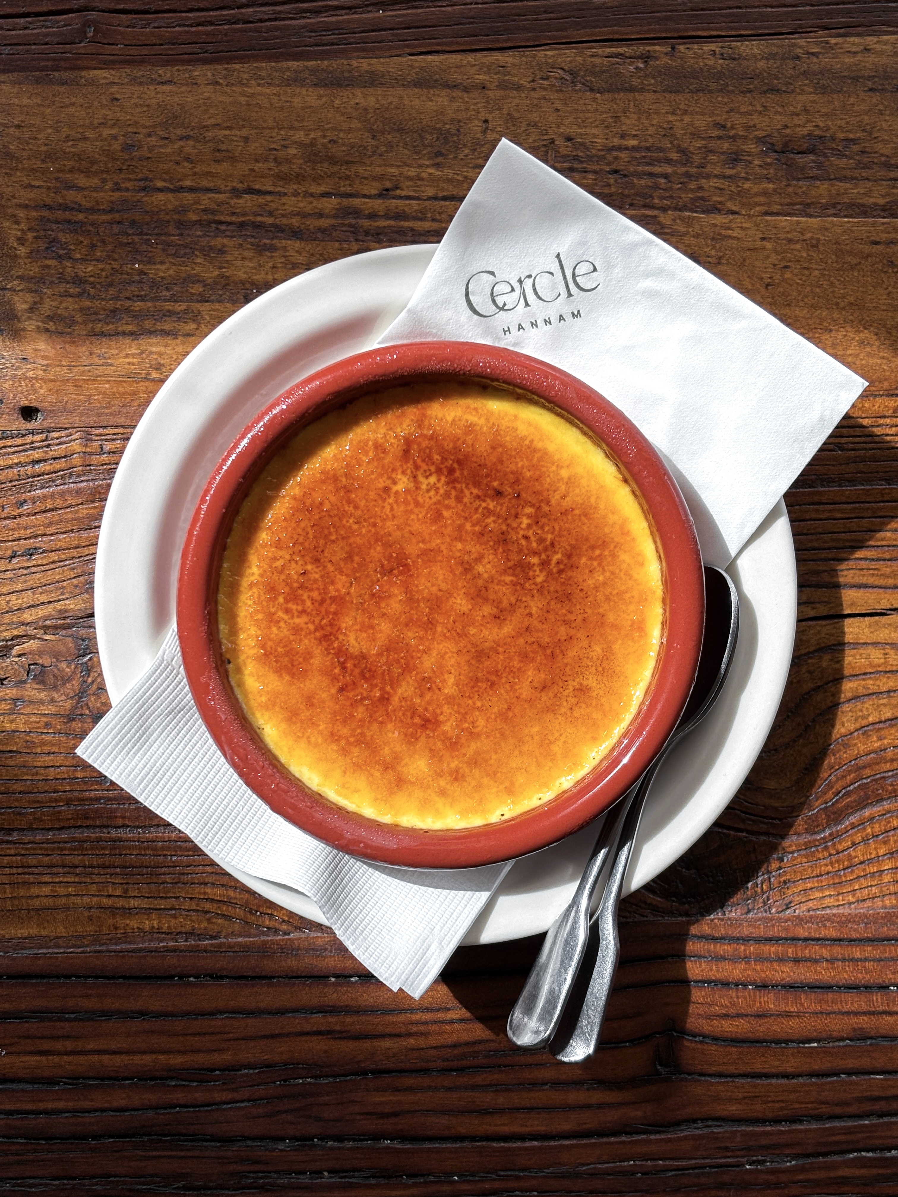 A top-down view of a crème brûlée in a terracotta-colored bowl on a wooden table at Cercle Hannam.