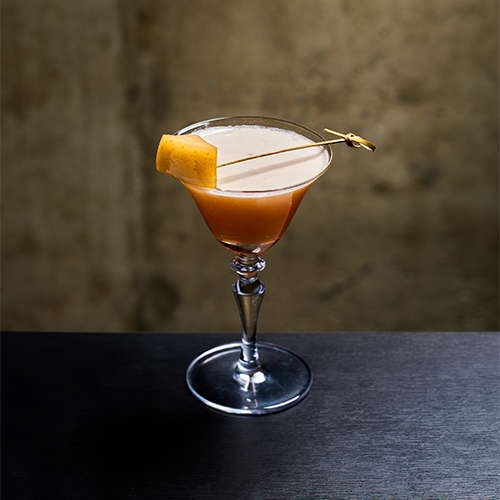 An orange cocktail in a martini glass with a gold garnish skewer against a dark, textured wall.